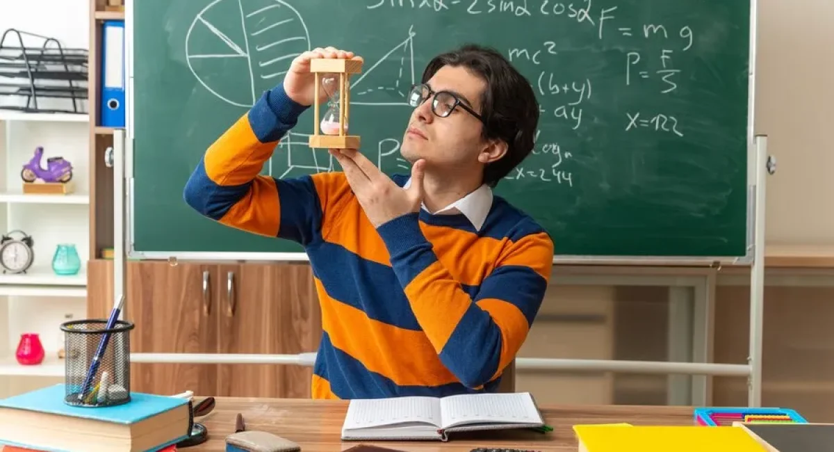 serious-young-caucasian-geometry-teacher-wearing-glasses-sitting-desk-with-school-tools-classroom-holding-hourglass-vertically-looking-it_141793-105472_11zon